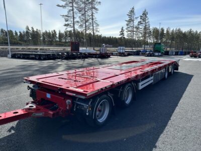 Röd bodvagn med stor flakyta snett framifrån framför större väg.