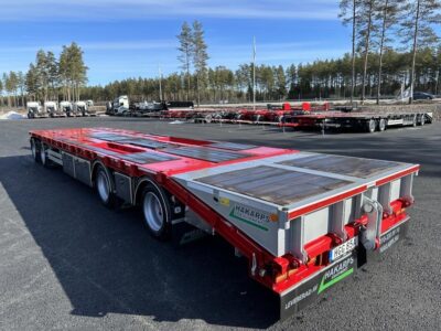 Röd bodvagn med stor flakyta snett bakifrån framför väg och fordon.