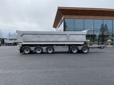 Grå dumpervagn med låsbar vändkrans från sidan framför byggnad.