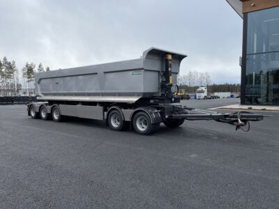 Grå dumpervagn med låsbar vändkrans snett framifrån med lastbil och byggnad bakom.